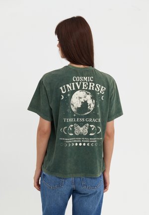Kvinde med langt brunt hår iført en grøn T-shirt med kosmiske månefaser, en sommerfugl og tekst om universet på ryggen.