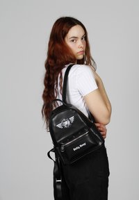 Mochila de cuero sintético negro con herrajes plateados, que presenta un parche de logo, tachuelas y un bolsillo frontal con cremallera. Correas de hombro ajustables.