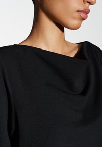 Indumento nero con collo alto in tessuto liscio e testurizzato. La ripresa ravvicinata mostra il collo drappeggiato e il dettaglio delle spalle sagomate, evidenziando un design elegante.