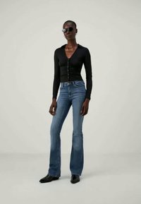 Top negro de manga larga con botones, jeans acampanados azules y gafas de sol negras. La modelo lleva unos zapatos negros elegantes y puntiagudos. Textura del tejido suave.