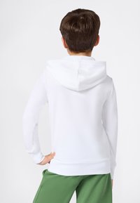 Felpa bianca con cappuccio regolabile con coulisse, tessuto morbido, maniche lunghe, orlo e polsini a costine, indossata con pantaloni verdi, vista da dietro.