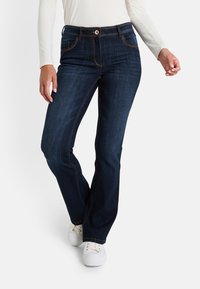 Mujer vestida con jeans bootcut azul oscuro, camiseta de manga larga blanca y zapatillas blancas, de pie con una pierna cruzada al frente.