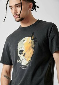 T-shirt en coton noir avec un graphique central de crâne en jaune et blanc, accompagné du texte "VIVRE/TRAVAILLER/CRÉER" en dessous. Col rond, manches courtes.