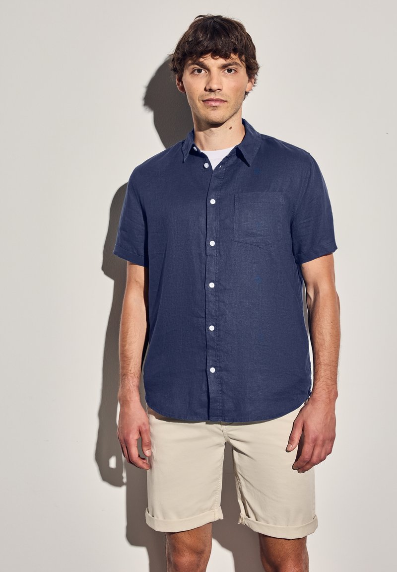 Krátký rukáv, tmavě modrá košile na knoflíky z lnu s náprsní kapsou, doplněná o vyhrnuté světlé béžové šortky. Hladká textura.