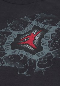 Μαύρο βαμβακερό t-shirt με γραφική εκτύπωση ενός κόκκινου σχήματος που σπάει μέσα από σχέδιο γκρι-ραγισμένου τοίχου. Μαλακή υφή.