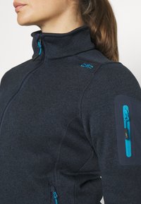 Kvinde iført en mørk marineblå fleece-jakke med lynlås og blå lynlåsgreb samt en lynlåslomme på venstre overarm.