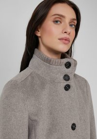 Vrouw met lang, donker haar die een lichtgrijze wollen jas draagt met een hoge ribbelkraag en vier grote zwarte knopen.