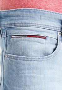 Ljust blå denimjeans bakficka med röd, vit och svart randig detalj, bärs med en röd melerad skjorta.