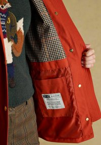 Czerwona kurtka z wierzchu w kratkę, złotymi detalami i wewnętrzną kieszenią. Zawiera białą metkę z logo i instrukcjami pielęgnacji.