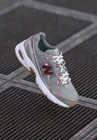 Baskets de sport grises avec tige en mesh, accents rouges, lacets blancs et semelle en caoutchouc chunky. Présente un logo sur le côté et le talon.