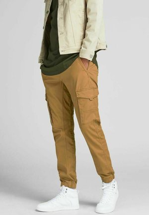Tan cargobroeken met elastische boorden, voorzien van meerdere zakken en een relaxte fit, gecombineerd met een lang groene shirt en witte sneakers.