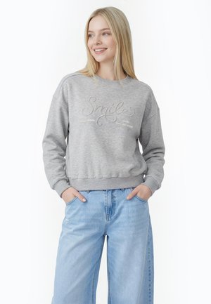 Ung kvinde med blondt hår iført en grå sweatshirt med teksten "Smile" og lyseblå bukser med vide ben, hænderne i lommerne, smilende let.