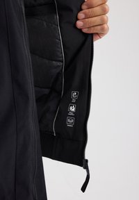 Schwarze gepolsterte Jacke mit einem glatten Innenmaterial, ausgestattet mit einem Reißverschluss und Symbolen für winddichte, wasserabweisende und gepolsterte Eigenschaften.