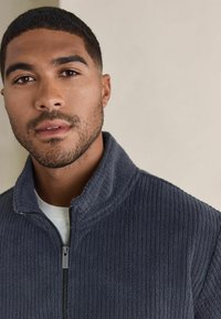 Dark gray zip-up fleece jacket with ribbed texture, high collar, and metal zipper. Worn over a white t-shirt.