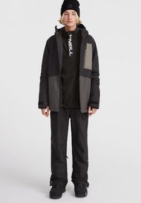 Chaqueta de nieve negra y gris con múltiples bolsillos, combinada con pantalones negros y un gorro negro. Cuenta con un cierre frontal y un logo en el pecho.