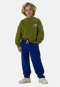 Sweatshirt vert avec logo, joggings bleus et baskets blanches. La tenue comporte des poignets côtelés et un cordon de serrage à la taille.