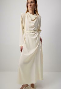 Naine kannab pikka kreemja satsiga kleiti pikkade varrukatega, drapeeritud kaelusega, vöödiga taljes ning kreemjas kõrge kontsaga sildiga kingi.