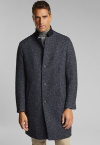 Uomo che indossa un soprabito herringbone grigio scuro con colletto e bottoni neri, dolcevita grigia e pantaloni beige su uno sfondo grigio chiaro.