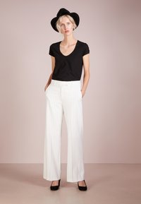 Schwarzes V-Ausschnitt-T-Shirt, weit geschnittene weiße Pinstripe-Hose und schwarze High-Heels. Model trägt einen schwarzen Hut. Neutraler Hintergrund.