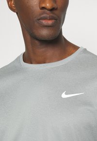 Nærbillede af en person iført en lyst grå skjorte med et hvidt Nike swoosh-logo på venstre brystområde.