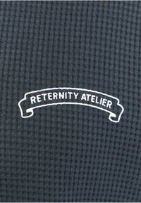 Tissu texturé sombre avec une bannière blanche incurvée contenant le texte « RETERNITY ATELIER » en lettres majuscules.