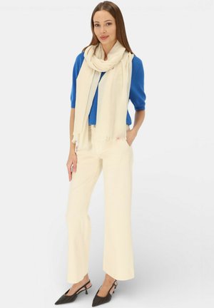Kobieta w kremowych spodniach typu szerokie nogawki, kremowym szaliku, niebieskim sweterku z krótkim rękawem i czarnych sandałkach na obcasie, stojąca z jedną ręką w kieszeni.