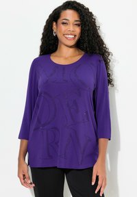 Violetti paita, jossa on kolme neljästä hihasta, ja suuri, kimalteisilla kivillä koristeltu teksti. Pehmeä kangas, jossa on rento istuvuus.