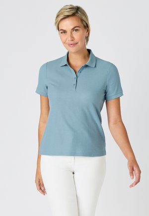 Mujer con cabello rubio corto que lleva una camiseta tipo polo de manga corta azul claro y pantalones blancos, de pie contra un fondo liso.