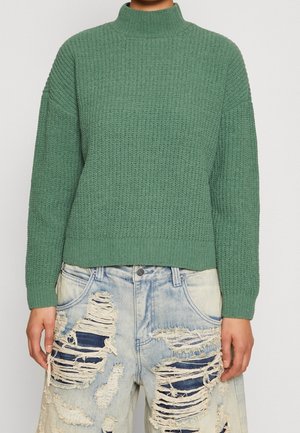 Personne portant un pull vert en maille à col montant et un jean bleu clair fortement déchiré.