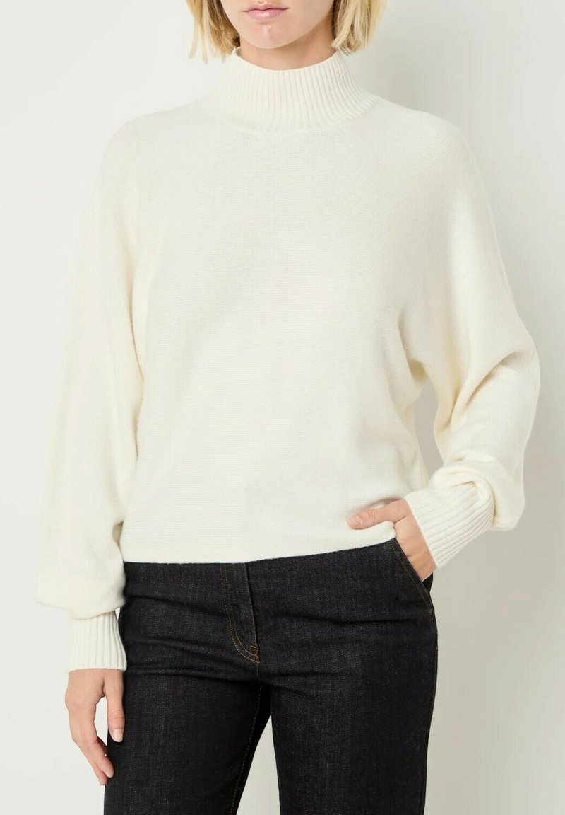 Maglione bianco lavorato a maglia con colletto alto a costine, spalle scese e polsini a costine. Abbinato a jeans in denim scuro.