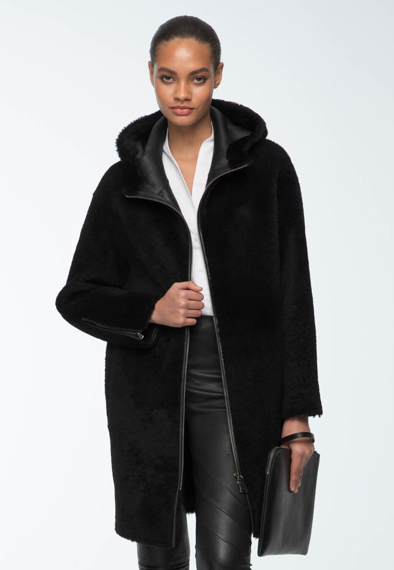 Cappotto in shearling nero con cappuccio, caratterizzato da un colletto in pelle nera a contrasto e zip, abbinato a pantaloni in pelle nera e una pochette.