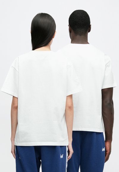 Dvije osobe stoje jedna uz drugu okrenute leđima, nose obične bijele majice i plave hlače s bijelim logotipima na lijevom bedrima.