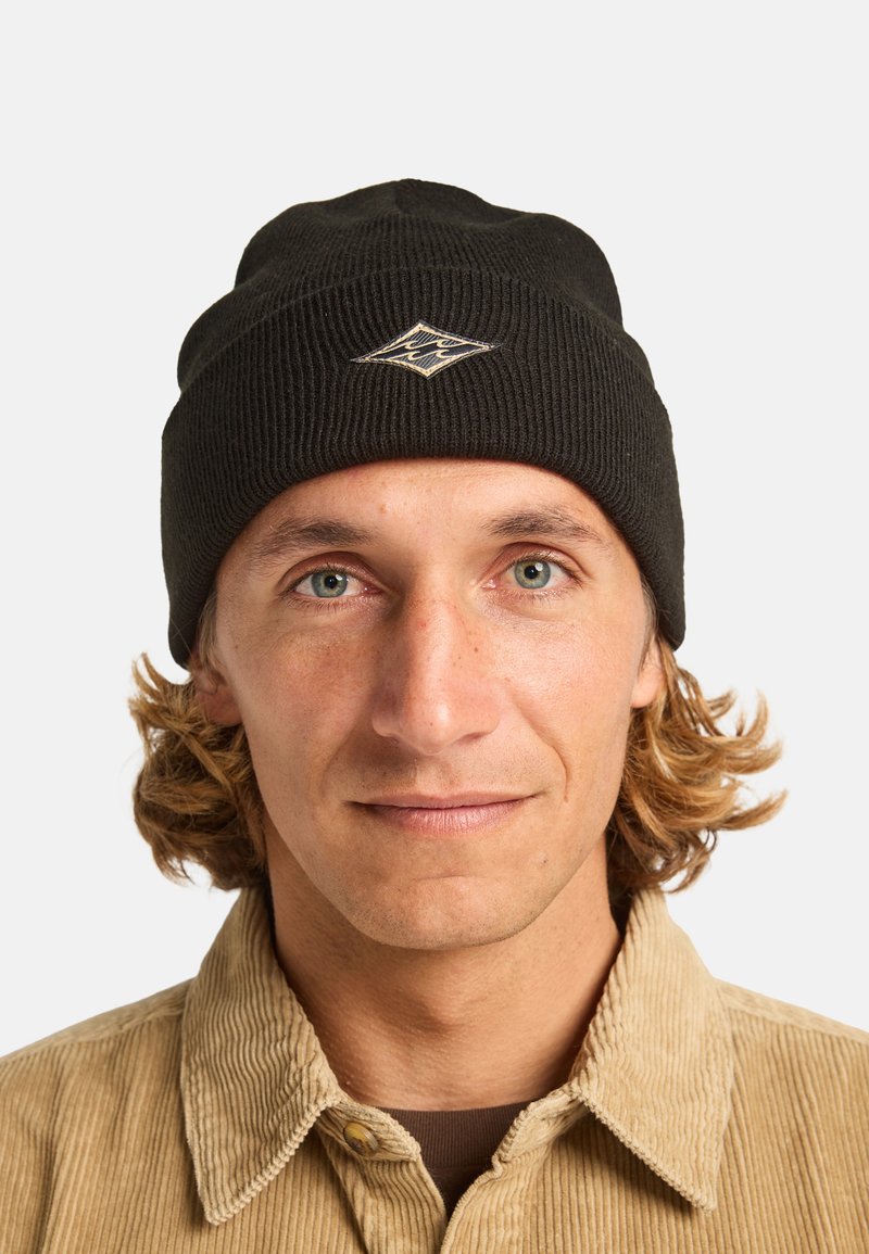 Beanie nero a coste con un logo a forma di diamante di colore contrastante sul davanti, indossato da una persona con capelli castano chiaro.