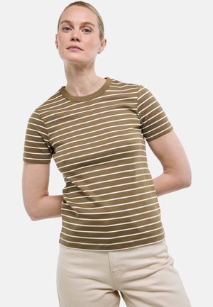 Femme aux cheveux blonds portant un chemisier à manches courtes rayé marron et blanc et un pantalon beige, debout les mains derrière le dos.