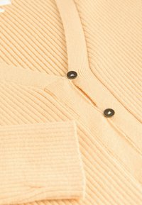 Ribbed, vaalean persikanvärinen neuletakki, jossa on V-kaula-aukko, kaksi mustaa nappia ja pehmeä pinta. Sisältää yhden avoimen taskun hihaassa.