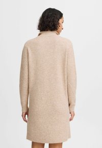 Brede gebreide jurk in zacht beige met een hoge kraag, lange mouwen en een gestructureerde afwerking. Beschikt over een knielengte en een geribbelde zoom.