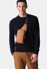 Brown leather sling bag with a smooth texture, featuring a zippered compartment and an adjustable black strap. Worn across the chest.