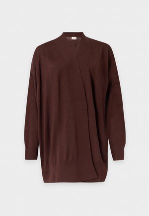 Femme debout avec une main dans la poche, portant un pull crème, un cardigan marron, un jean bleu et des chaussures à talons beiges sur fond blanc.