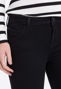 Czarne dżinsy o dopasowanym kroju, z pięcioma kieszeniami i subtelnym przeszyciem. Pas jest wyposażony w zapięcie na guzik i zamek błyskawiczny.