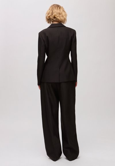Personne blonde de dos portant un blazer noir cintré et un pantalon noir ample sur un fond gris uni.