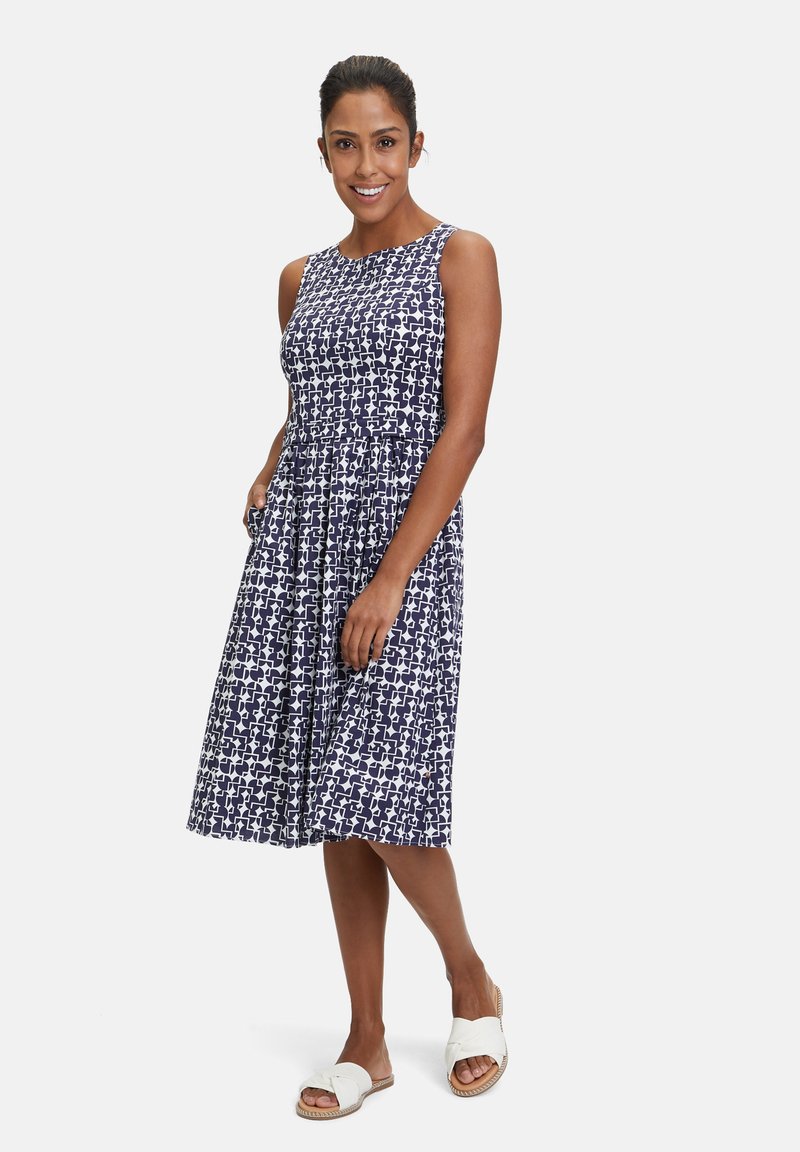 Marineblaue ärmellose Kleid mit einem weißen abstrakten Muster, tailliertem Oberteil, schwingendem Rock und Taschen. Getragen mit weißen Sandalen.