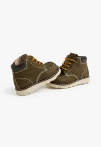 Bottines en cuir marron avec des lacets jaunes, des coutures blanches, des accents noirs et une semelle épaisse crème. Elles présentent une texture lisse et un bout arrondi.