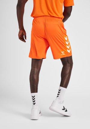Oranje sportshorts met een elastische tailleband, voorzien van een wit logo en chevronpatroon aan de zijkant. Gdragend met witte sokken en schoenen.