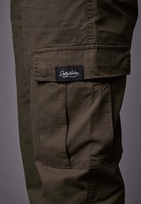Olivgrüne Cargo-Hosen aus einem strukturierten Stoff, mit seitlichen Taschen und einem schwarzen Etikett mit weißem Text "Dolly Noire" auf der linken Tasche.