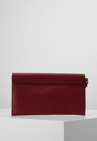 Mørkerød læder clutch-pung med klaplukning, præsenteret på en hvid pedestalsokkel mod en enkel lys grå baggrund.