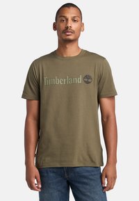 Camiseta de algodón verde oliva con el logo de "Timberland" y un gráfico de árbol en verde más oscuro. Cuello redondo clásico y mangas cortas.