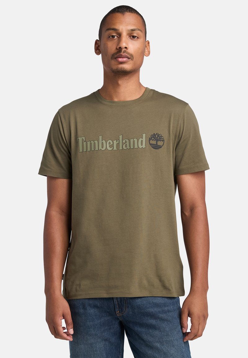 Camiseta de algodón verde oliva con el logo de "Timberland" y un gráfico de árbol en verde más oscuro. Cuello redondo clásico y mangas cortas.