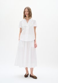 White short-sleeve blouse with a V-neck and pleat detail, paired with a white midi skirt. Black and brown leopard-print flats.
