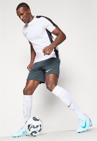 T-shirt atlético branco com painel lateral preto, calções verdes escuros, meias brancas e botas de futebol azul claro. Inclui uma bola de futebol com um padrão.