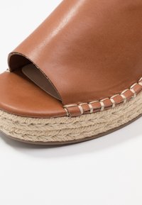 Sandale compensée ouverte en cuir marron avec une plateforme en jute tressé. Présente des détails cousus et une texture lisse sur la partie supérieure.